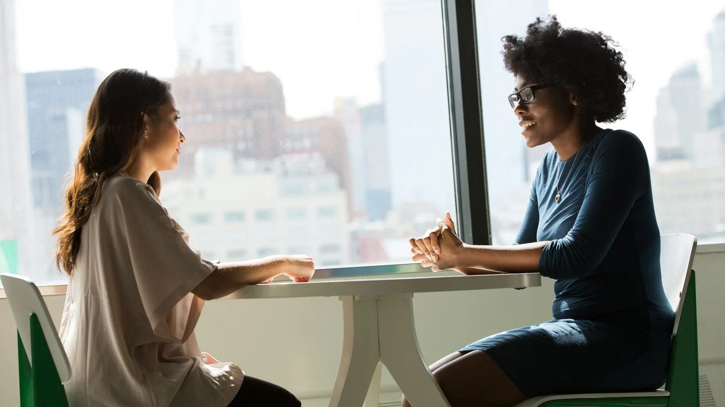 Two colleagues have a conversation at a table beside a window.
