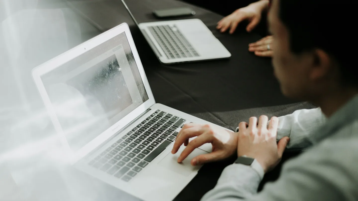 A person works on a laptop beside colleagues at a shared desk.