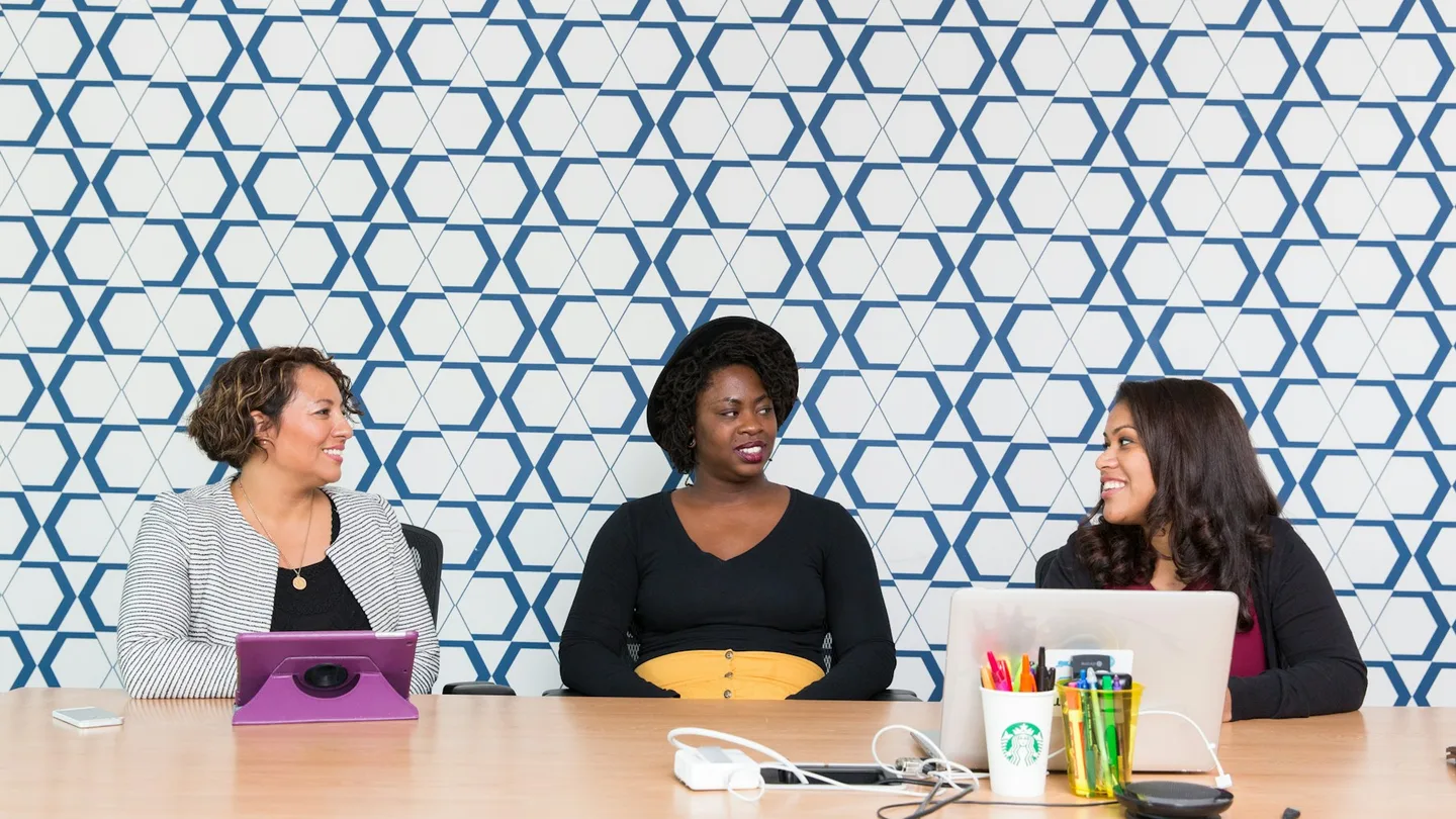 Three professionals discuss a project with a laptop and tablet at a meeting table.