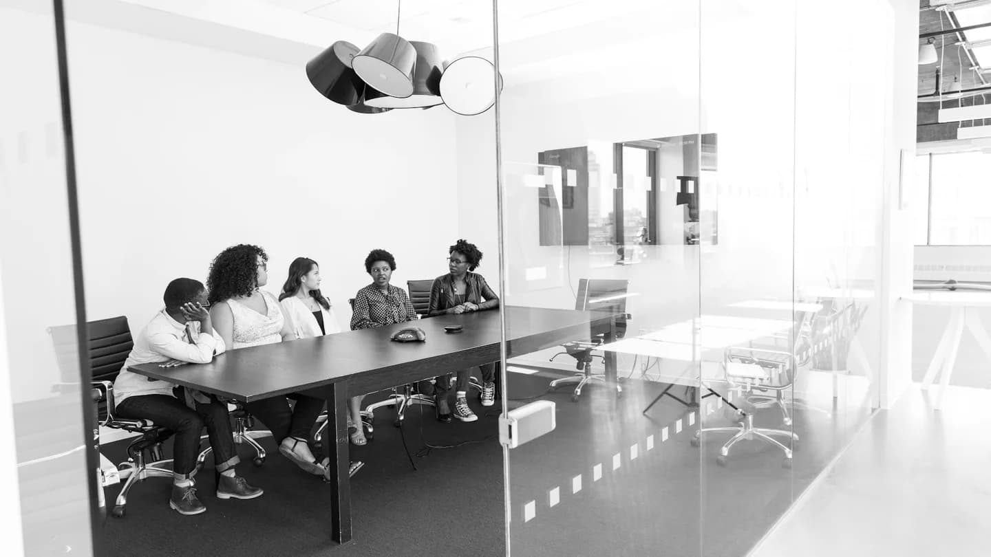 A team meets around a conference table in a bright office, photographed in black and white.