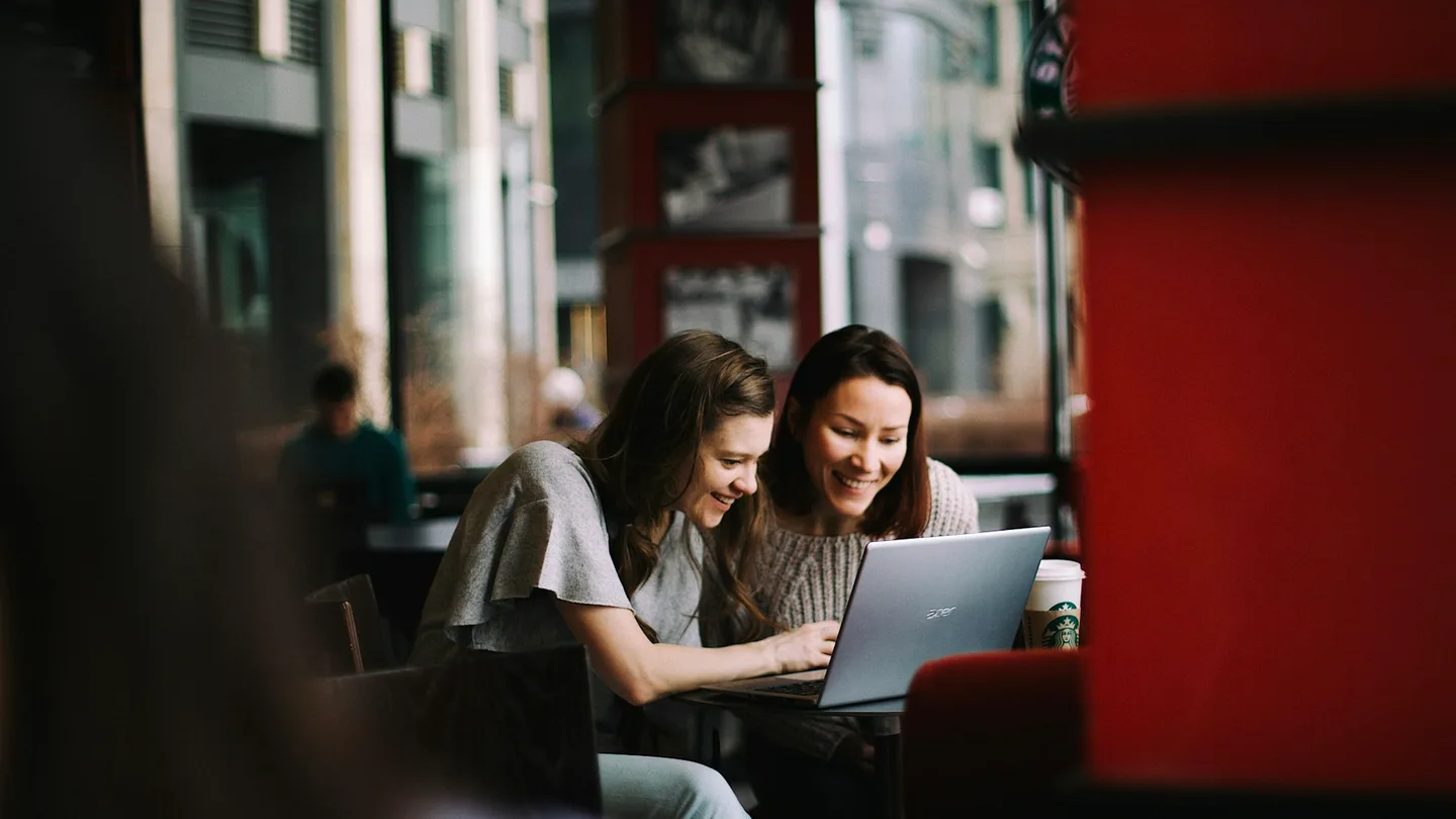 Two women review a laptop screen together in a cafe.