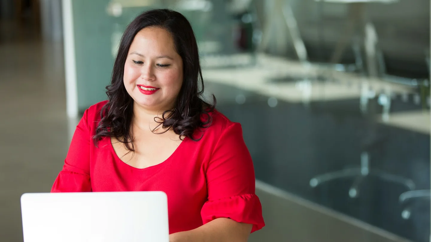 A woman in a red blouse works on a laptop in a bright office.