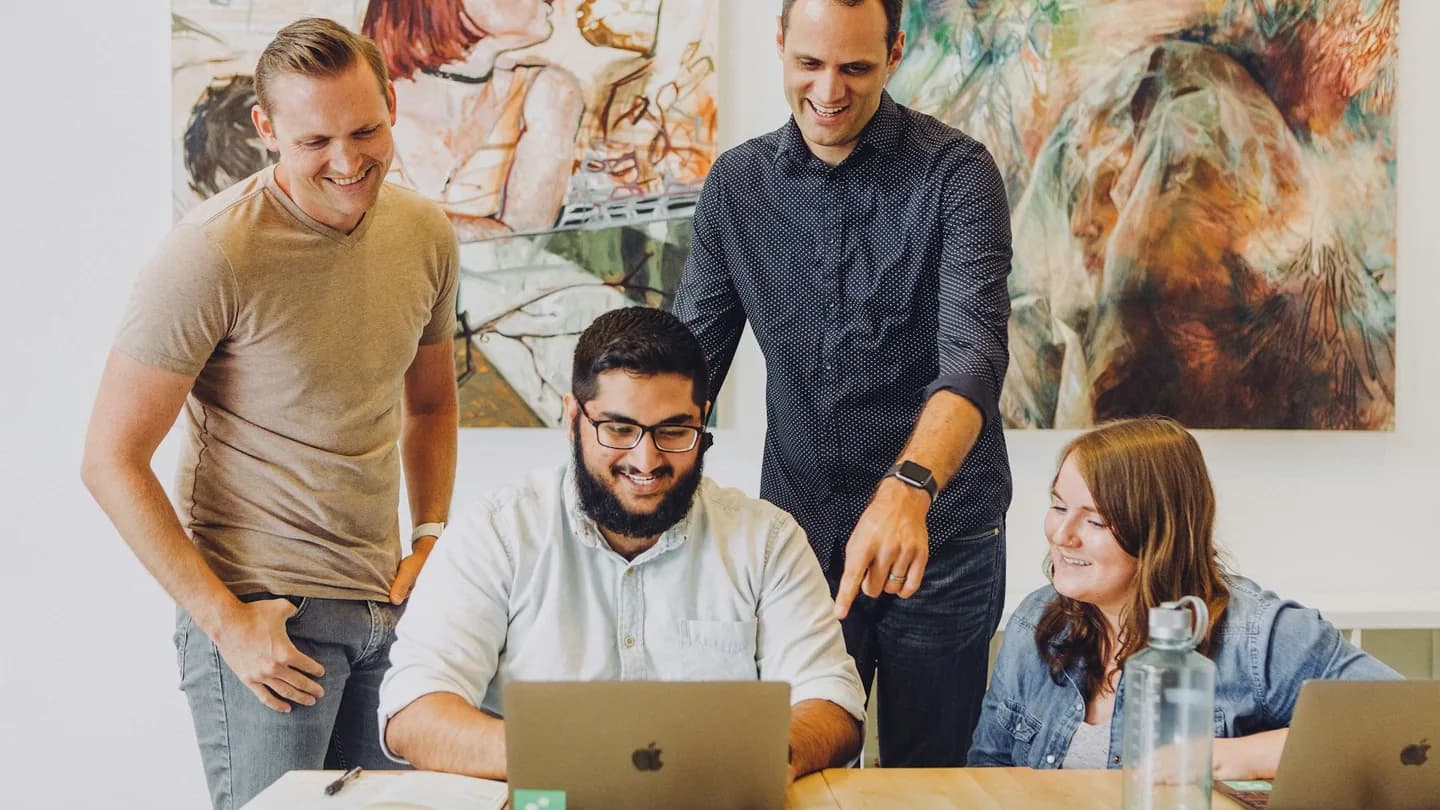 Four colleagues gather around a laptop at a wooden meeting table.