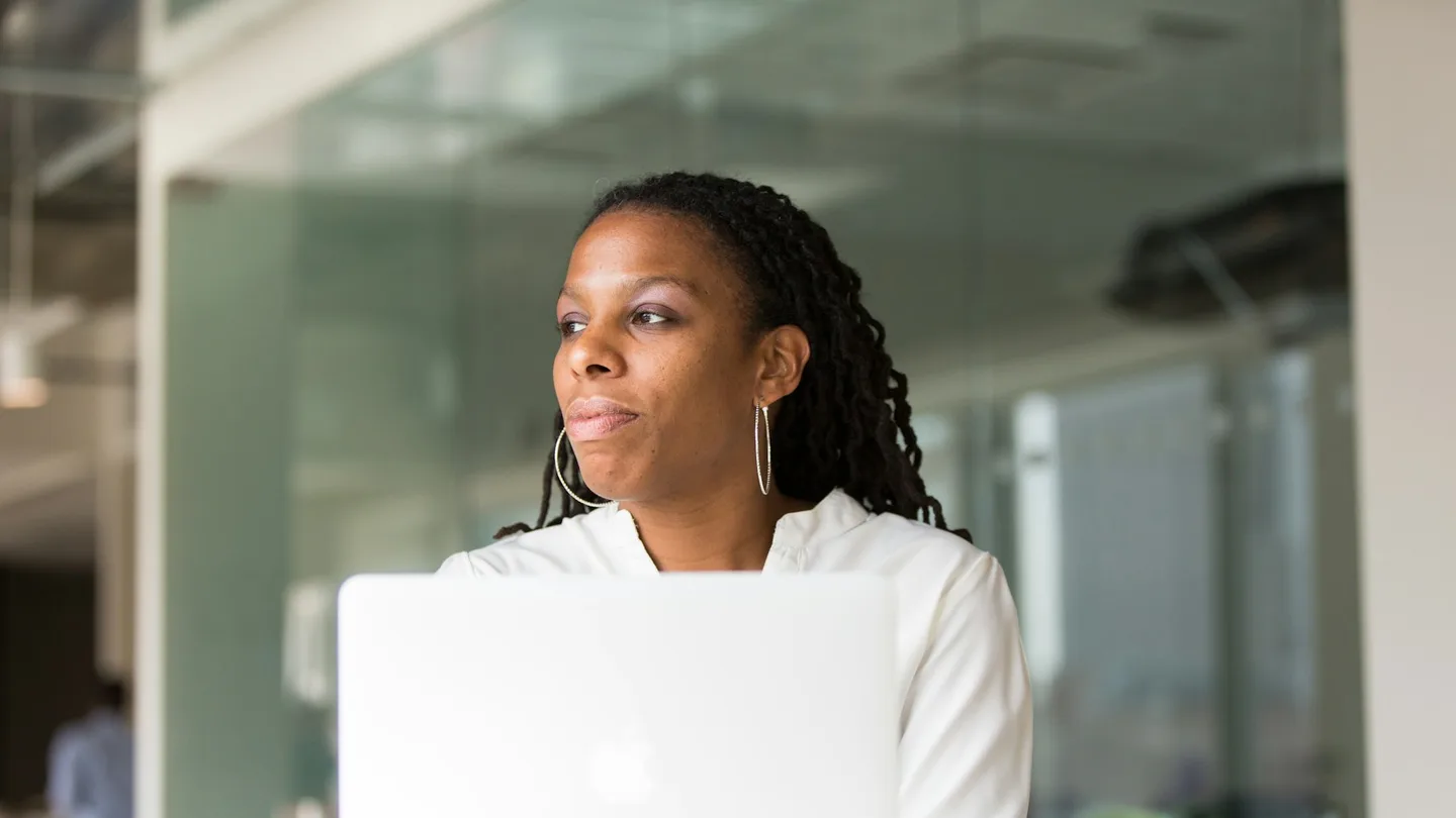 A woman pauses thoughtfully while working at her laptop.